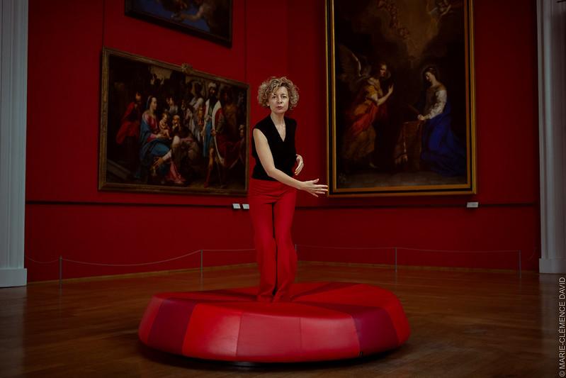Bernadette Gruson en pantalon rouge et haut noir sans manches.
Debout sur un grand pouf rond et rouge, dans une des salles du musée des Beaux-Arts de Lille.
Derrière elle, un mur rouge en angle avec d'immenses peintures de l'époque Renaissance.