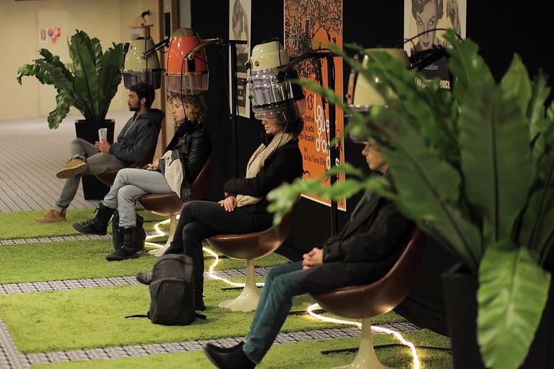 4 personnes assises dans des sièges dorés à pied blanc, sous des casques de permanente, écoutant les textes de Bernadette Gruson.