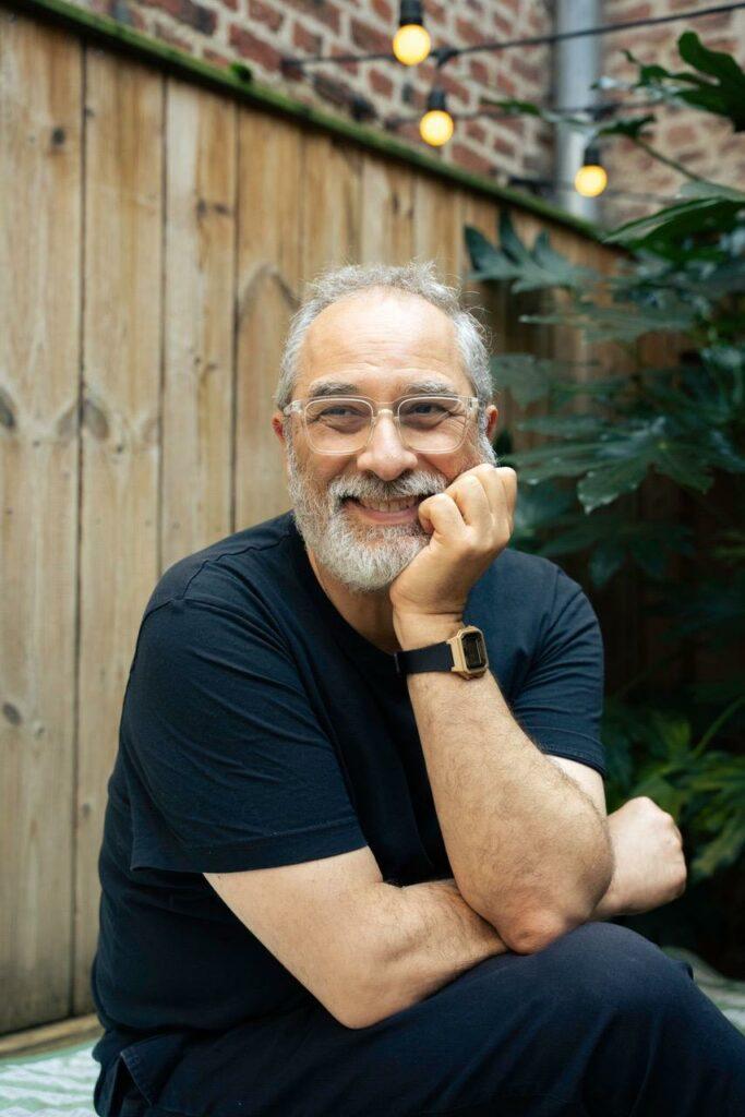 Mario Alonso, assis.
Il porte un T-shirt et un pantalon sombre, noir ou bleu très foncé.
Le visage dans la main gauche, souriant.
En arrière-plan, une palissade en bois clair, un mur de brique et des plantes.