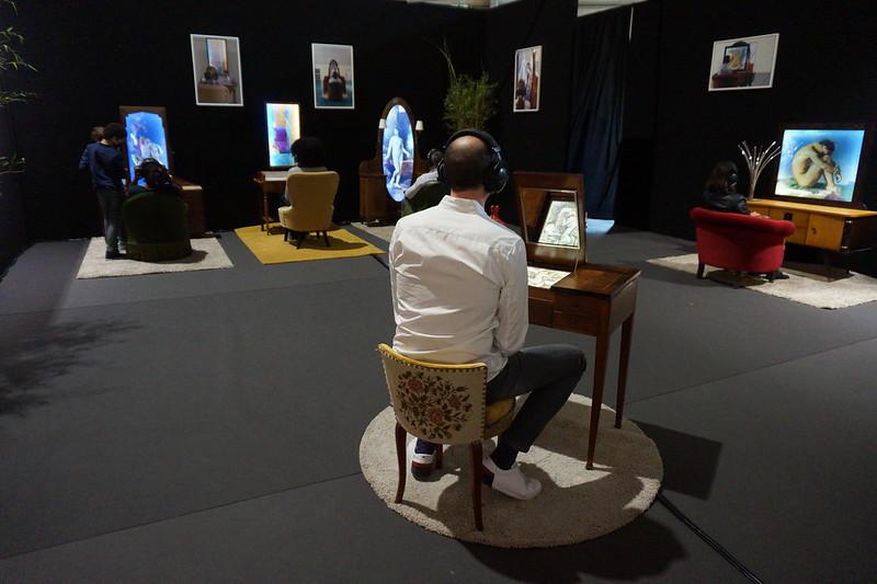 Plusieurs coiffeuses/tables de toilette avec des sièges.
Chaque miroir/écran affiche un tableau de nu.
Des personnes sont assises sur les sièges, casque sur les oreilles, et écoutent les textes de Bernadette Gruson.