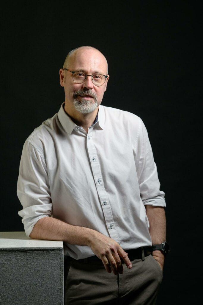 Portrait vertical de Jean-Christophe Piot.
Il est debout, appuyé sur un bloc balnc.
Il porte un pantalon gris et une chemise blanche, et une paire de lunettes.