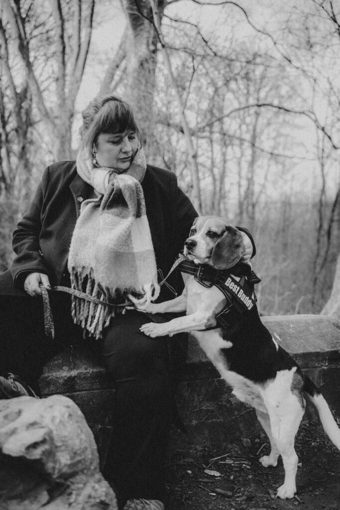 Photo en noir et blanc.

Sandrine Goeyvaerts, long manteau noir et écharpe. Elle est à l'extérieur, assise sur un parapet en pierre.
Elle tient en laisse son chien, Quinoa, un beagle, qui est dressé sur ses pattes arrière et les pattes avant sur sa cuisse.
