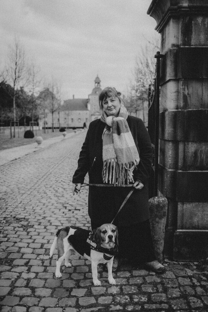 Photo en noir et blanc.

Sandrine Goeyvaerts debout contre un pilier en pierre, dans une rue pavée.
Elle porte un long manteau noir et une écharpe. Elle tient en laisse son chien, Quinoa, un beagle.