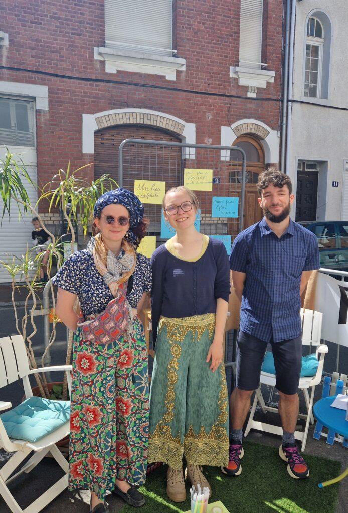Trois personnes debout.
Une femme en pantalon à fleurs, haut fleurs et turban bleu.
Une femme en pantalon vert et doré, haut bleu.
Une gomme en short et chemise bleue.
