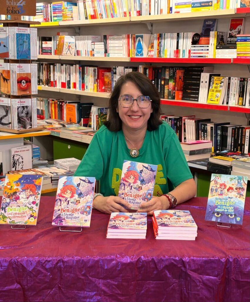Blandine Butelle est assise devant une table où sont présentés les 3 premiers tomes des aventures de Bénigne.
Elle porte un haut vert.
Derrière elle, des étagères de librairie, couverts de livres.